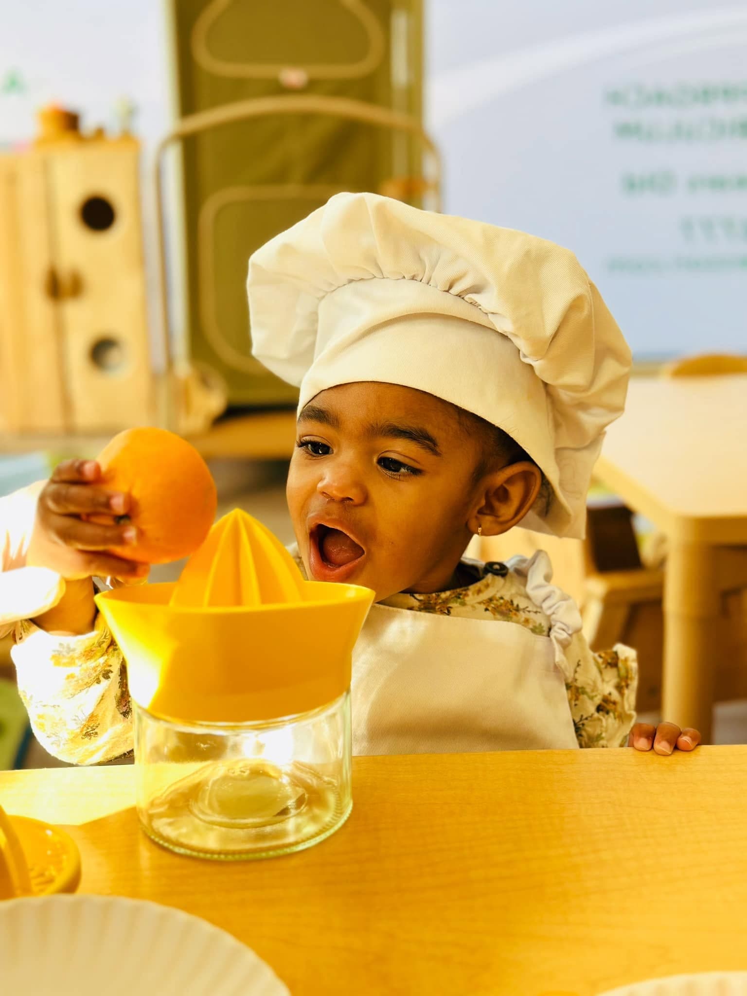 Toddler in a chef hat and apron joyfully juicing an orange during a cooking activity at Kinder Prep