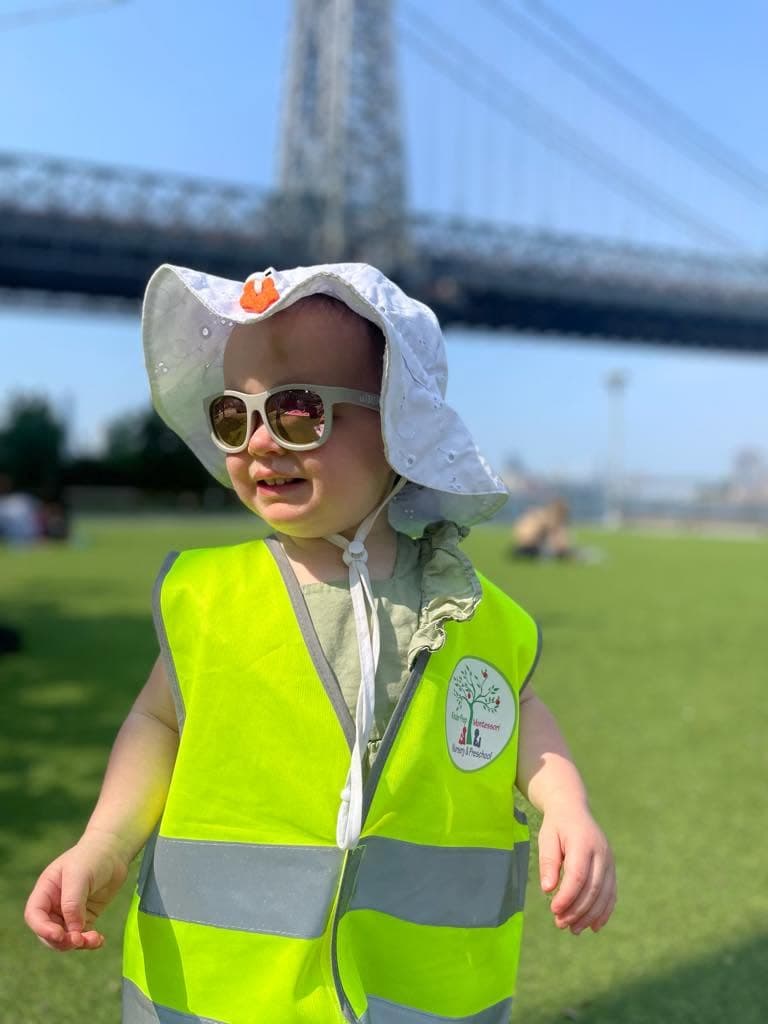 Toddler in a sun hat and safety vest enjoying an outdoor field trip by the Williamsburg Bridge